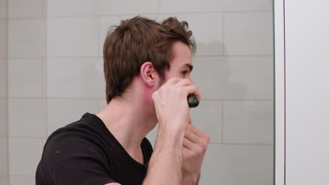 young man trimming and shaving his own beard in front of mirror in bathroom