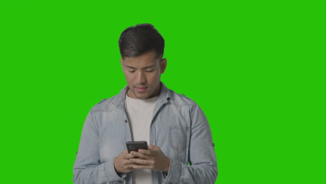 Studio-Shot-Of-Casually-Dressed-Young-Man-Online-Swiping-On-Mobile-Phone-Against-Green-Screen-