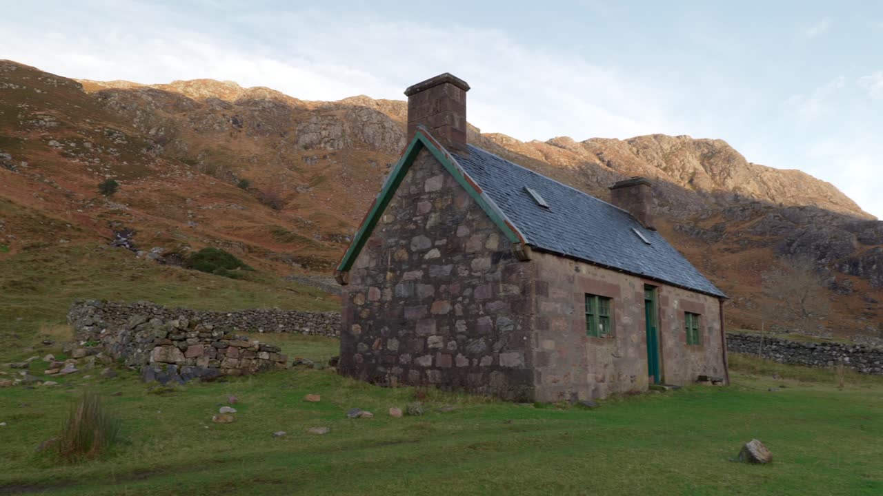 una panorámica derecha lenta revela un antiguo edificio de piedra en la base de una montaña en una cañada remota en la escocia rural