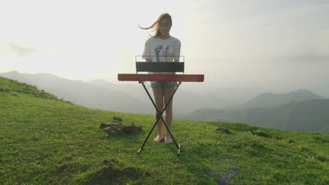 Drone-Acercándose-A-Una-Joven-Músico-Tocando-El-Teclado-Del-Piano-En-La-Naturaleza-Pura-Al-Atardecer-Montañas-Paisaje-Del-Valle-Verde