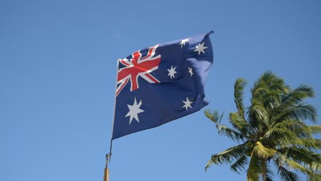 Hermosa-Bandera-Australiana-En-Cámara-Lenta-Con-Viento-Y-Hermosa-Palmera-Detrás-En-La-Playa-En-Verano