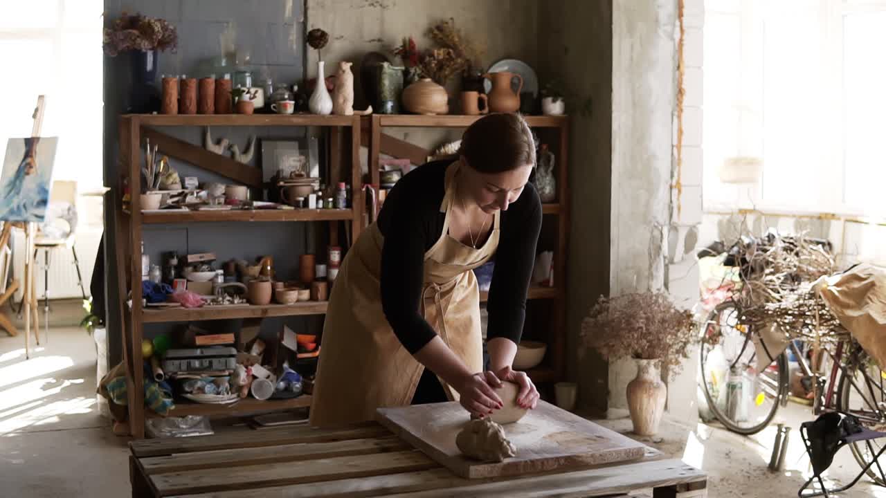 Portrait of female potter wearing beige apron putting clay piece on worktop and then starting kneading it with her hands. Pottery products on shelf behind