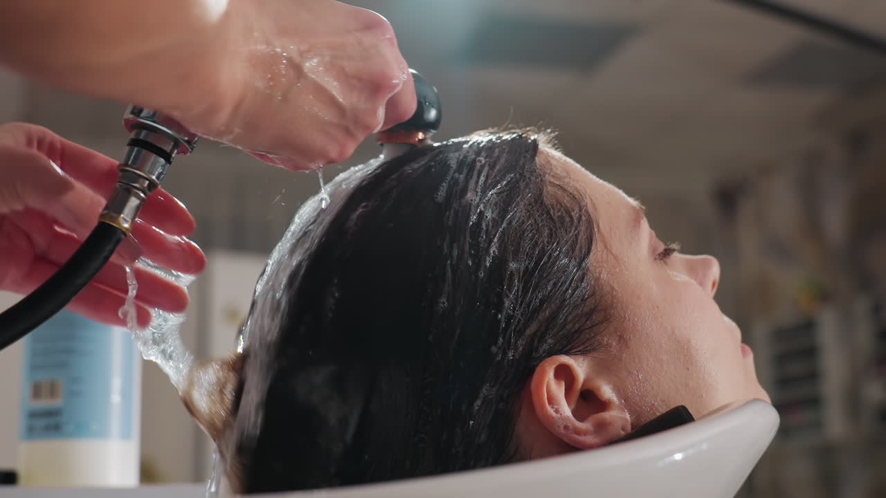 미용실 싱크대에서 흐르는 물에 머리를 감는 모습 클로즈업, 두피에서 헹궈지는 거품 샴푸, 머리카락 가닥을 따라 움직이는 집중된 손길, 선명한 빛과 배경의 제품 병