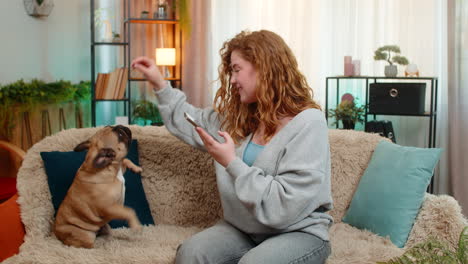 young woman taking photo of cute pug dog on home sofa smiling and creating cheerful pet content