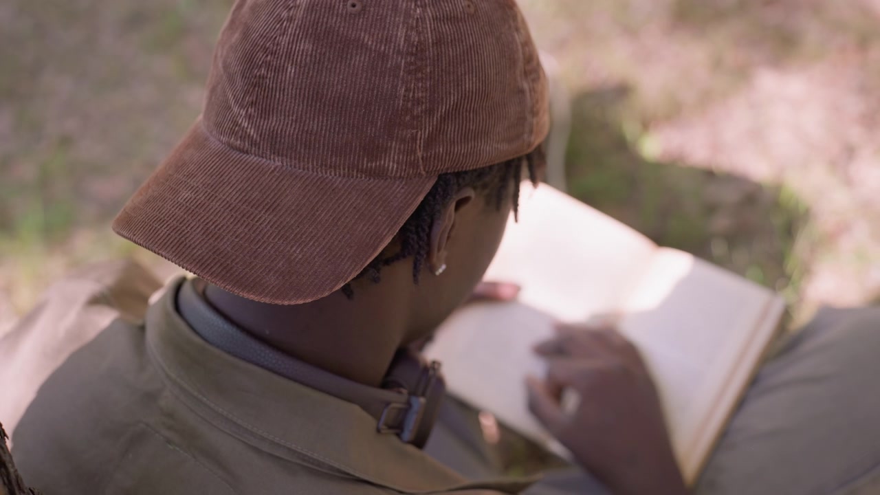 Close up of young woman reading open book outdoors with sunlight casting soft shadows on her face and page. Woman wears brown corduroy cap green jacket details visible including straps