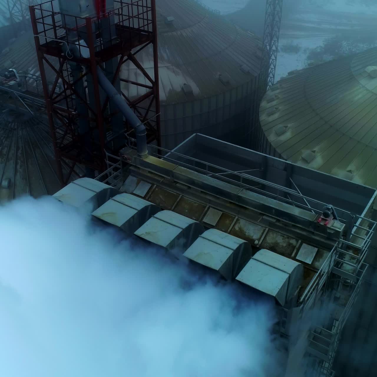 Thick white smoke coming from the pipes at grain elevator. Aerial shot of the metal tanks silo on grey foggy day