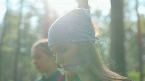 close-up of cousins walking through lush forest, one cousin wearing blue scarf and softly lit by sunlight, gazing downward as other cousin is softly blurred in background, light filters through trees