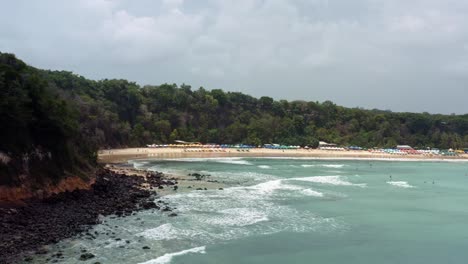 Right-trucking-aerial-drone-shot-of-the-famous-tropical-Madeiro-beach-near-Pipa,-Brazil-in-Rio-Grande-do-Norte-with-calm-waves-for-surfing,-colorful-umbrellas,-and-surrounded-by-large-exotic-cliffs