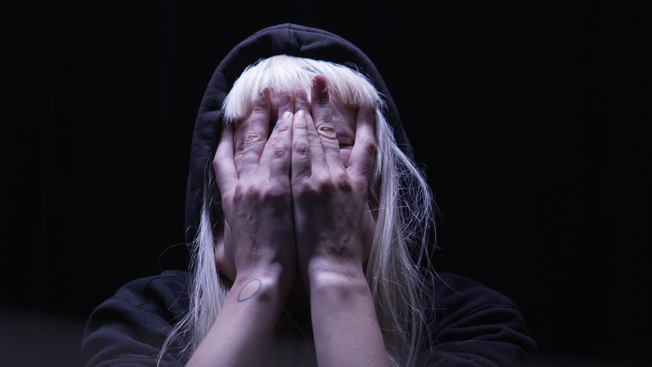 하나의 오버헤드 조명으로 조명된 희미한 방에서 두 손으로 얼굴을 문지르는 겁먹은 사람