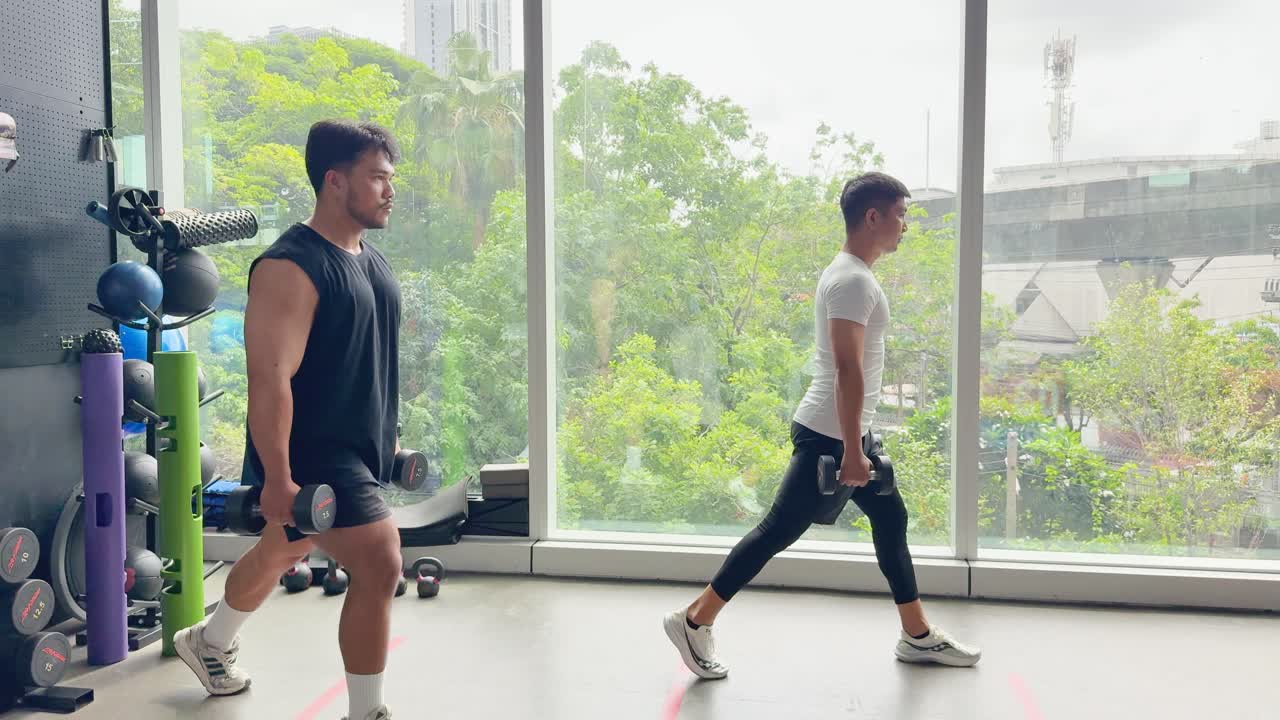Two men exercise with dumbbells, performing lunges in a well-lit gym with large windows
