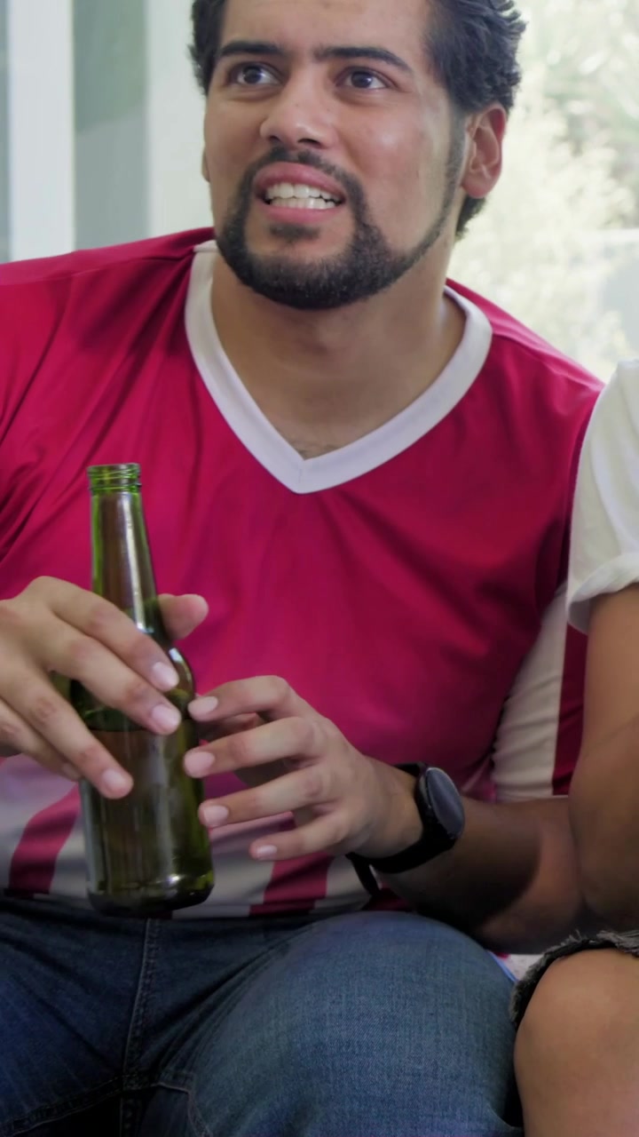 Vídeo vertical: Homem de camisa vermelha reagindo a um feed fora da tela no sofá, segurando uma garrafa verde.