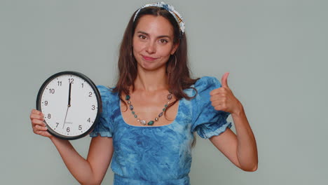 mujer bonita mostrando la hora en el reloj de la pared de la oficina, ok, pulgar hacia arriba, aprobar, señalando con el dedo a la cámara