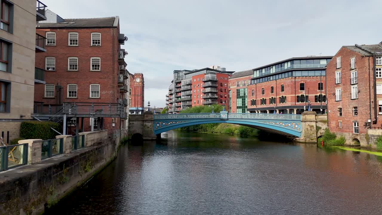 Camera moves forward along the River Aire toward a blue Victorian bridge, flanked by historic and modern buildings under daylight with overcast skies