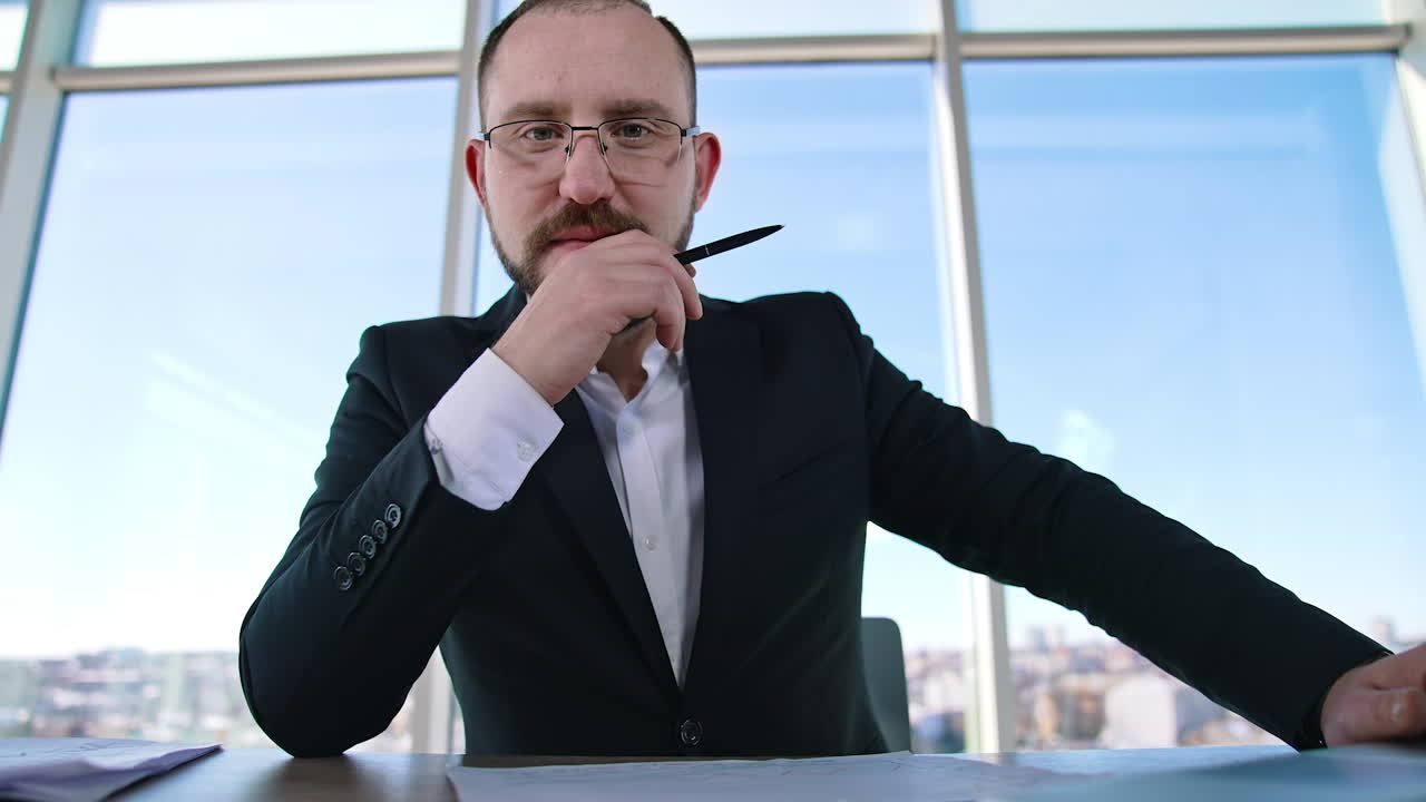 Portrait of positive businessman. Successful entrepreneur sitting at table and looking at camera. Bearded boss in glasses and elegant suit in office.