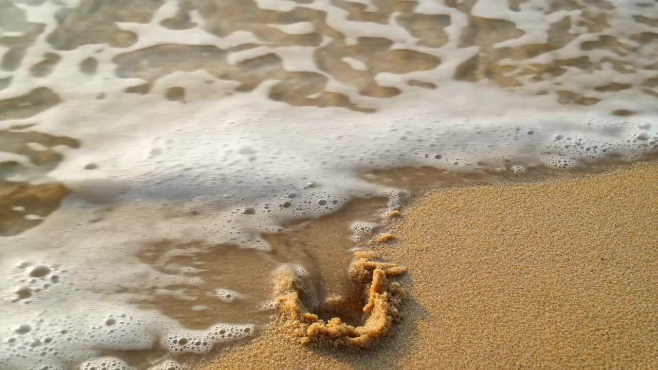 Scena ravvicinata della costa con schiuma di mare bianca che si muove sulla sabbia bagnata, mentre il bordo dell'onda principale entra dall'angolo superiore sinistro e si diffonde attraverso l'inquadratura. Un piccolo