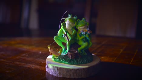 couple frog figurine on wooden presentation base with lights. male frog holding fishing rod while sitting next to his lovely girlfriend. close up