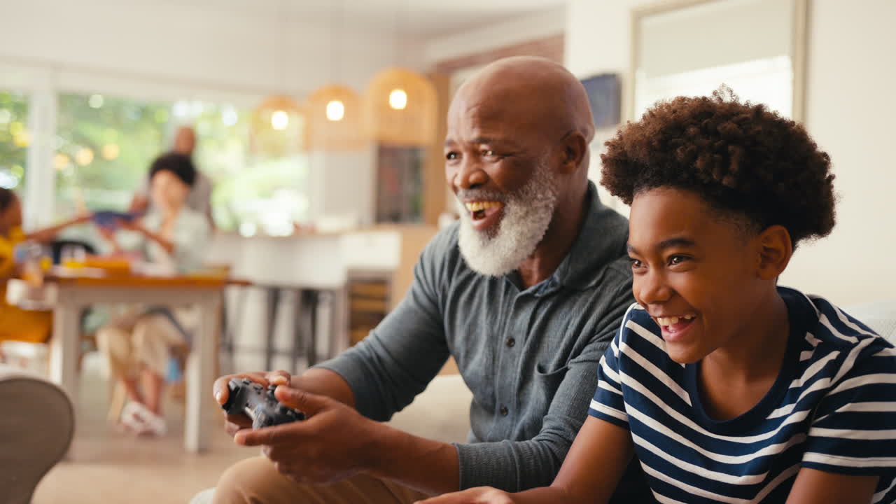 Grandfather And Grandson Sitting On Sofa At Home Playing Video Game Together