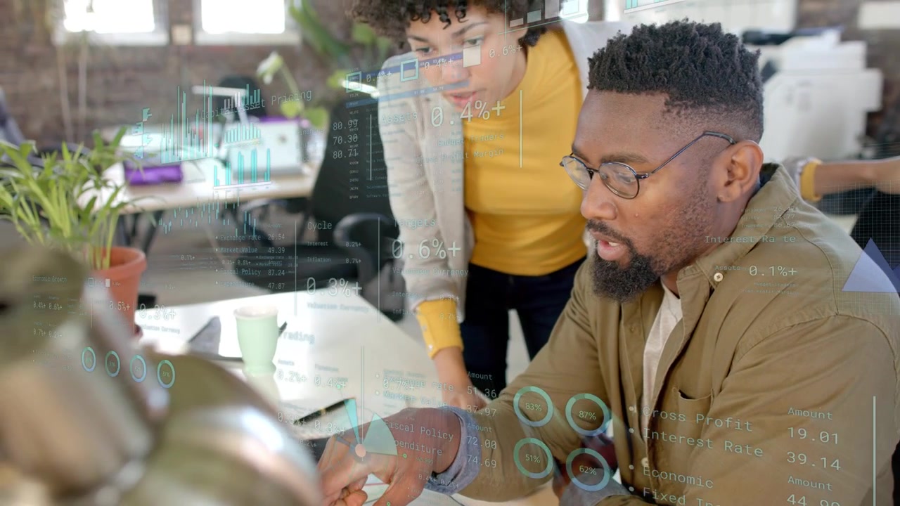 Standing woman pointing, seated man tapping keys, charts over laptop following taps analyzing data