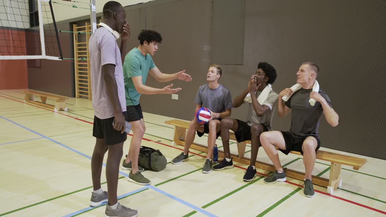 Ein diverses Männerteam sitzt auf einer Turnhallenbank; die Person im grünen T-Shirt beginnt ein Gespräch, hält einen Volleyball und bereitet sich auf das Spiel vor.