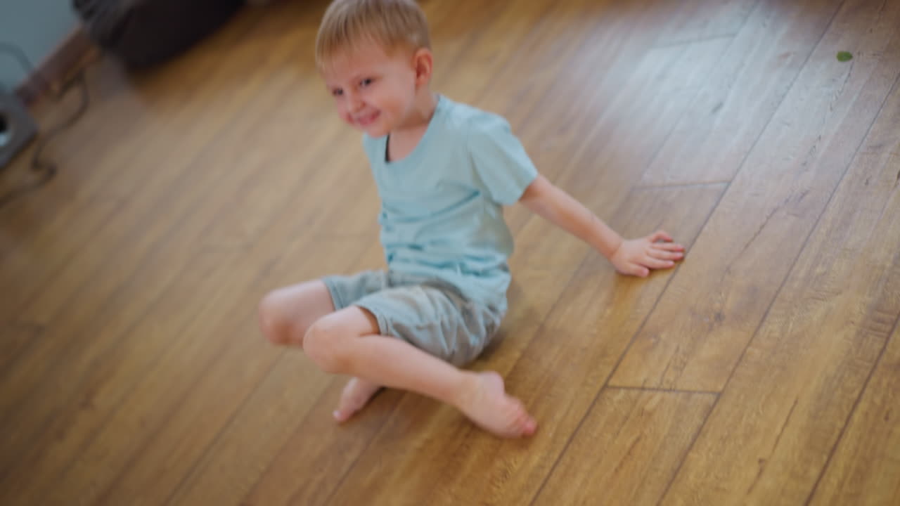 Bambino energico in pantaloncini gioca su un pavimento di legno in casa, colto a metà movimento mentre cade in avanti, esprimendo uno stile di vita attivo, divertimento, spensieratezza e gioia dell'infanzia attraverso un movimento giocoso spontaneo