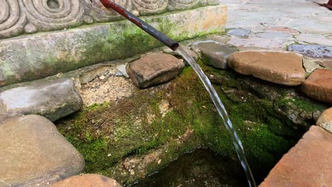 Close-up-View-at-Water-Flows-from-a-Metallic-Hose-to-a-Stone-and-Mossy-Well-the-Monastery-from-a-Cave---Ialomita