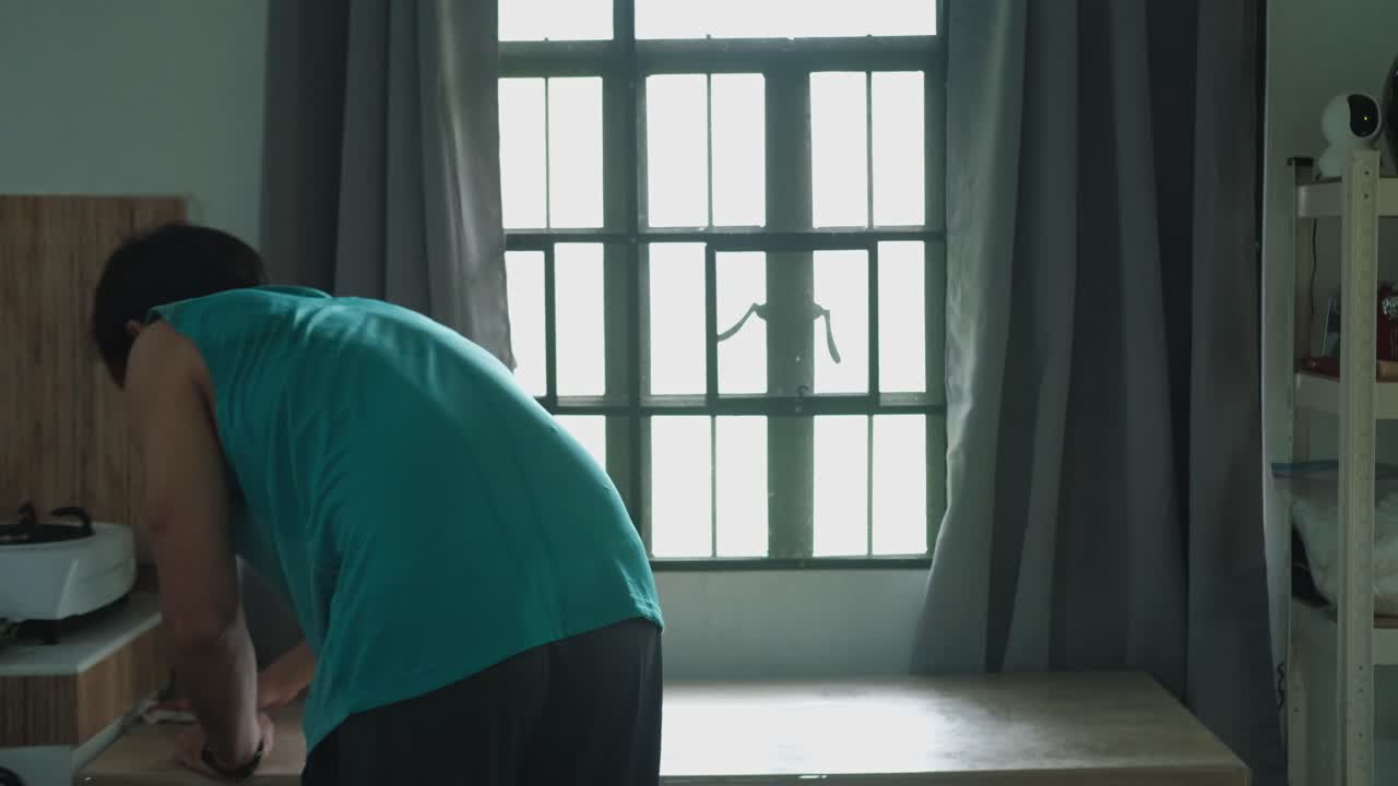 Rear static shot of young man cleaning kitchen table while facing aesthetic light from apartment window.