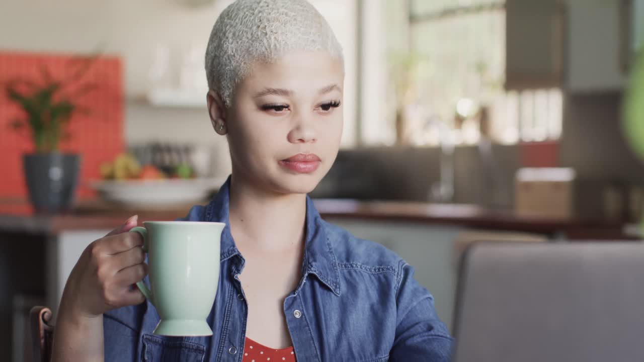 mujer biracial ocupada trabajando en casa con una computadora portátil y bebiendo café en cámara lenta