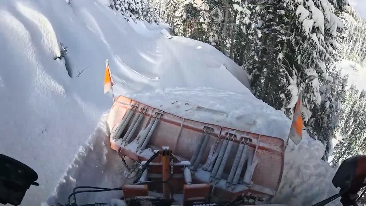 FPV or first person view from inside a snow plowing machine that is plowing and pushing snow if front of it in the mountains during a sunny day. The road ahead is covered with thick white fluffy snow.