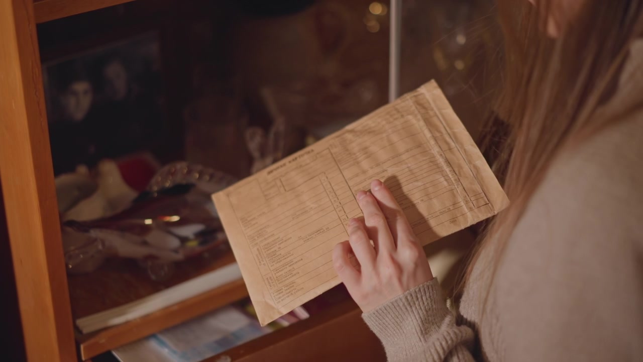 Una donna caucasica scopre una lettera scritta a mano in un cassetto di una vetrina; mani lente e delicate estraggono una busta fragile da un cassetto foderato di velluto tra ninnoli vintage, sotto una calda luce ambrata, con un primo piano della calligrafia.