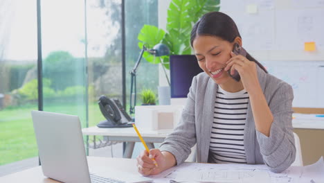 Arquitecta-Que-Trabaja-En-La-Oficina-Sentada-En-El-Escritorio-Hablando-Por-Teléfono-Móvil