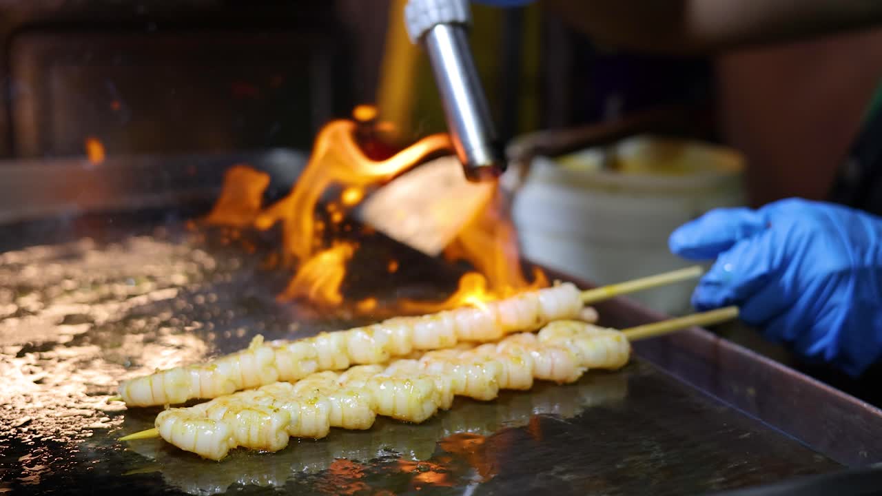 Sælger med handsker bruger en blæselampe til at grille rejespyd på en kogeplade, livlige flammer, street food om natten