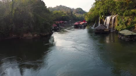 A-wonderful-4K-drone-shot-of-a-river-in-the-jungle-of-Thailand,-looking-at-a-small-floating-village-and-a-beautiful-tropical-waterfall-in-Sai-Yok-National-Park-in-Asia