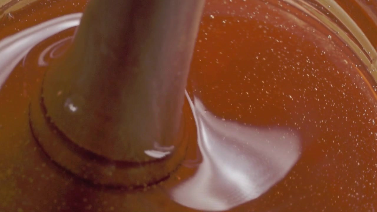 Macro close up of honey being stirred with a wooden dipper featuring golden color thick consistency glossy texture and slow flowing movement