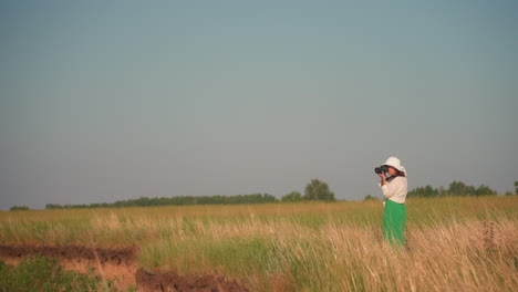 female photographer stands near cliff holding camera surrounded by tall grass in golden field under blue sky, capturing scenic outdoor landscape in peaceful countryside