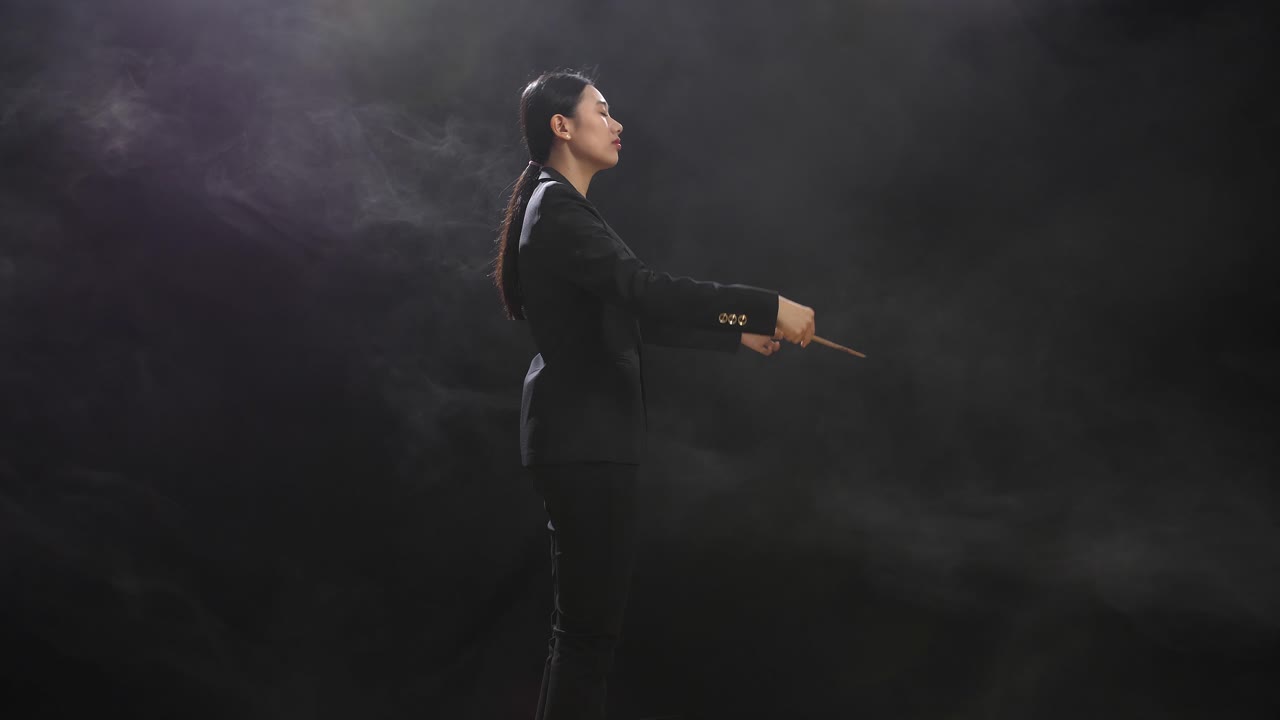 Vista laterale di una donna direttrice d'orchestra asiatica che tiene una bacchetta, chiude gli occhi e mostra un gesto in uno studio nero con nebbia.