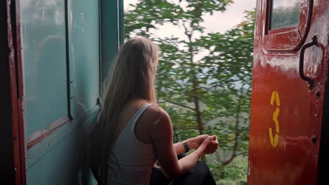 a woman sits at the open door of a moving train on the iconic ella to kandy route in sri lanka, calmly enjoying the stunning landscape of tea plantations, hills, and tropical forest.