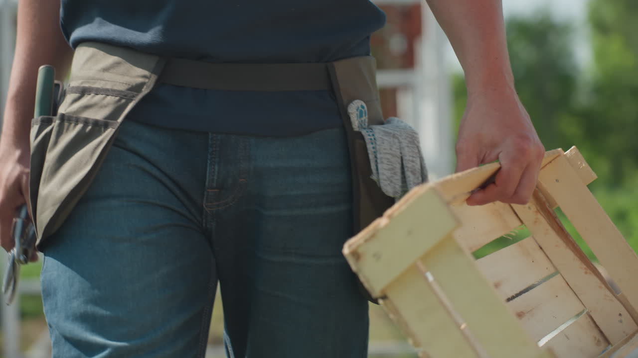 Close up worker carrying wooden crate while walking around garden plot wearing tool belt with gloves, sunlight on denim pants and greenery blurred in background