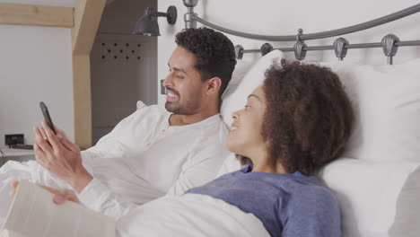 Couple-In-Bed-Wearing-Pyjamas-With-Woman-Reading-Book-And-Man-Taking-Selfie-On-Mobile-Phone