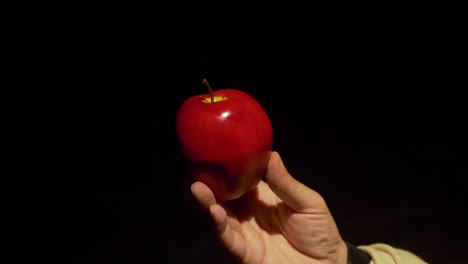 Eine-Weiße-Männerhand-Dreht-Langsam-Einen-Leuchtend-Roten-Apfel-Vor-Einem-Komplett-Schwarzen-Hintergrund