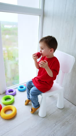 cute baby boy sits on a chair near the big window. toddler holds two apples and eats one of them. high angle view. vertical video.
