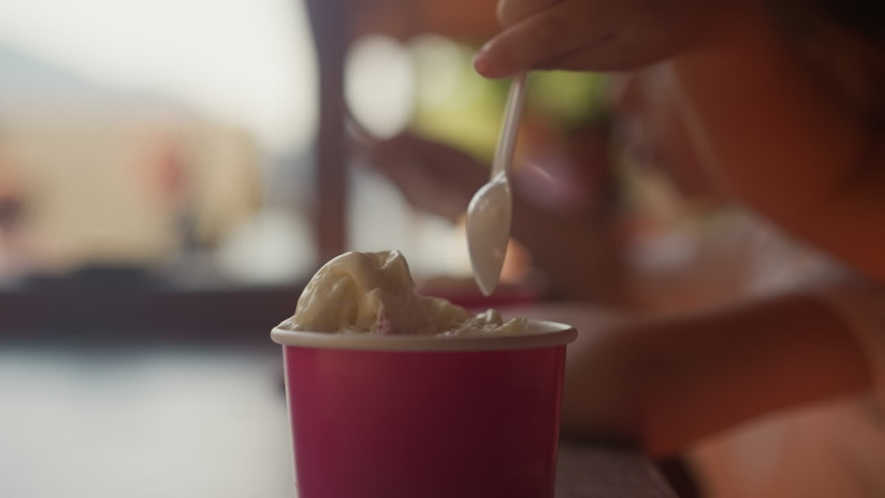 Little child picks ice cream with spoon from paper cup sitting at cafe table closeup. Toddler kid has gelato for dessert in canteen. Summer treats