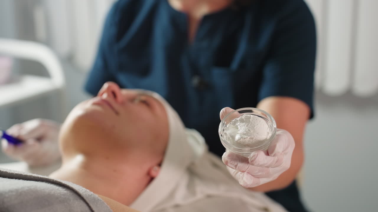 Beautician wearing protective gloves holds transparent silicone cup of white facial cream while using blue brush to apply mask onto client wrapped in head towel in bright serene spa room