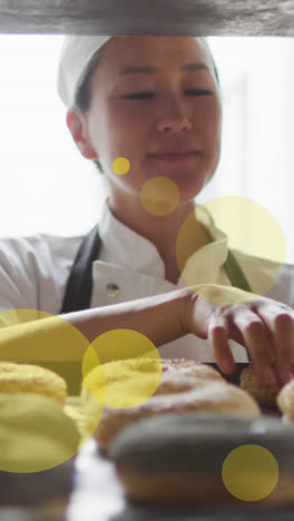 Animation-of-green-spots-over-asian-female-baker-preparing-donuts