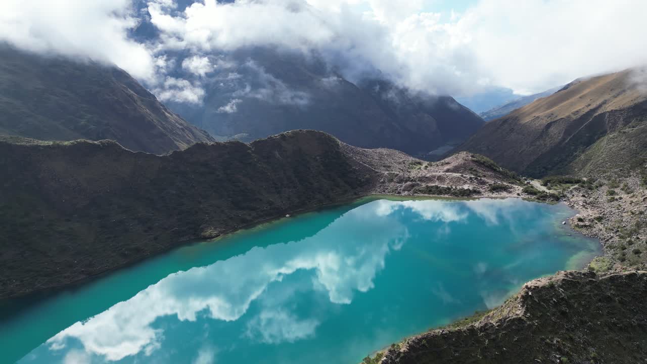 Aerial view of Humantay glacial lake in the Andes, showing turquoise water reflecting dramatic cloud patterns in clear, sunlit conditions