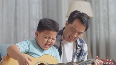 close up of asian father and son with expressive face enjoys singing and playing the guitar together on sofa at home