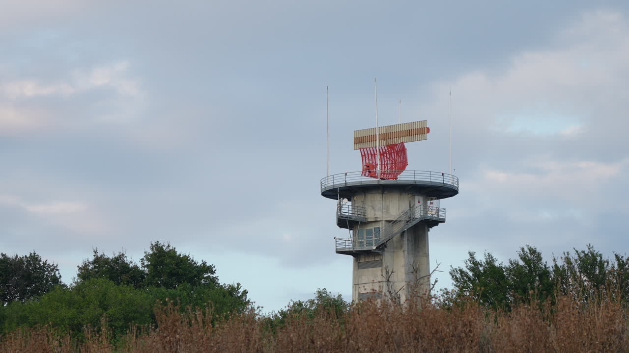 Concrete radar tower with spinning antenna located near forested area under evening sky