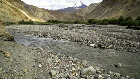 Tilt-up-shot-in-the-Himalayas,-as-a-river-flows-in-the-foreground,-with-the-snowy-peak,-Kongmaru-La-in-the-background