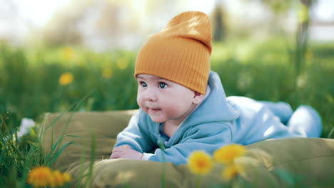 adorable baby boy in blue overall and orange cap in nature. cute child resting in the grass. blurred backdrop.