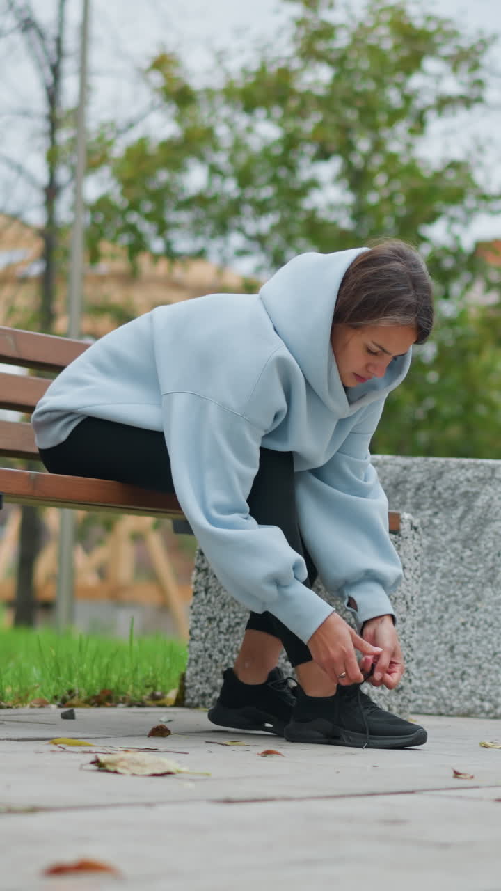 Girl sitting on concrete bench tightening shoelace in outdoor park, surrounded by trees and a pole in background, calm atmosphere, autumn setting, preparing for walk or exercise