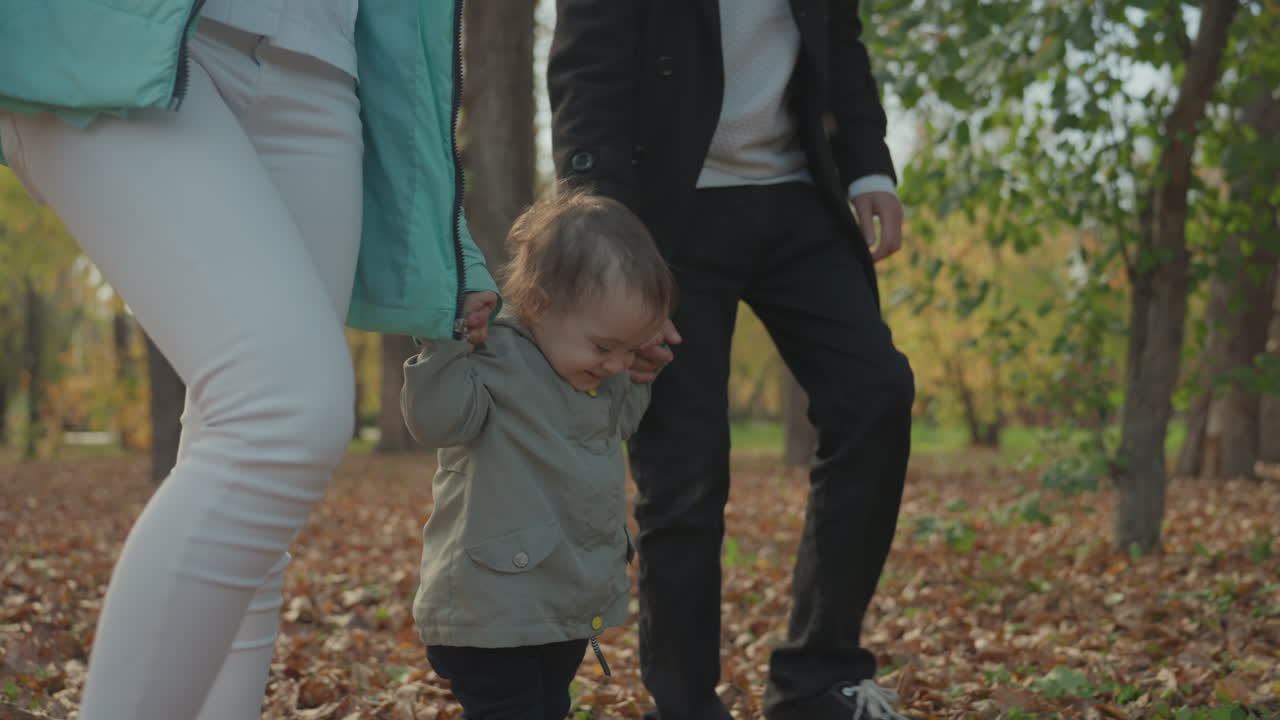 Genitori bianchi giocosi nel parco incoraggiano i primi passi del bambino, volti sorridenti e movimento vivace, foglie che volano, mani che sostengono il bambino, gita familiare attiva che cattura un'interazione gioiosa e il matrimonio